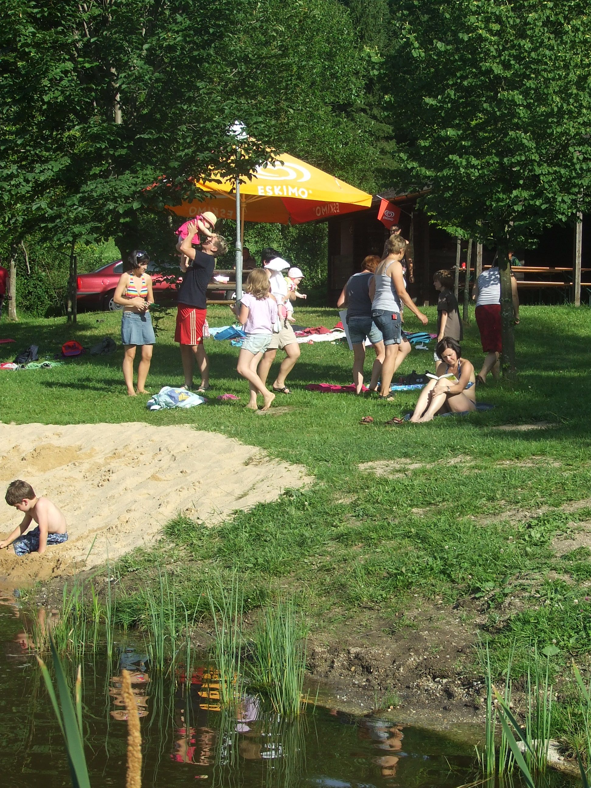 People relax by a natural bathing pond with a parasol and trees.