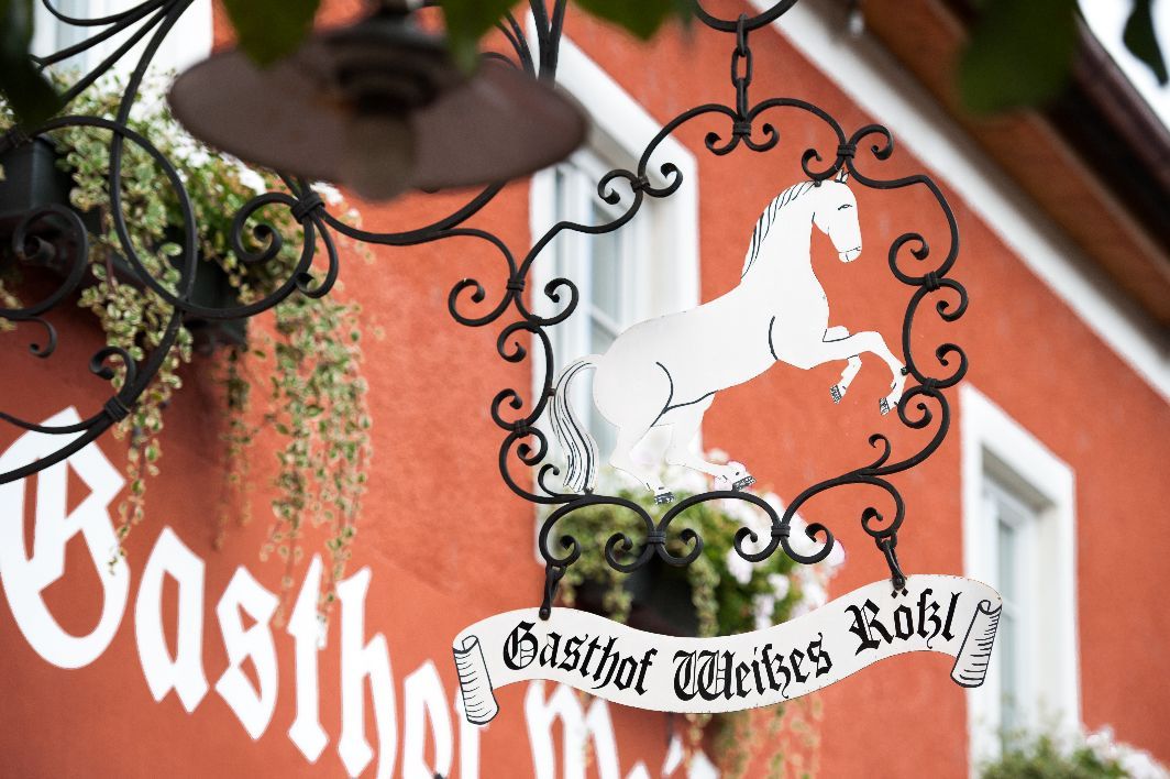 Metal sign with a white horse and the words 'Gasthof Weißes Rößl' in front of a red building.