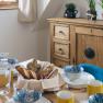 A laid breakfast table with bread rolls, crockery and yellow cups, a wooden cupboard with plants in the background.
