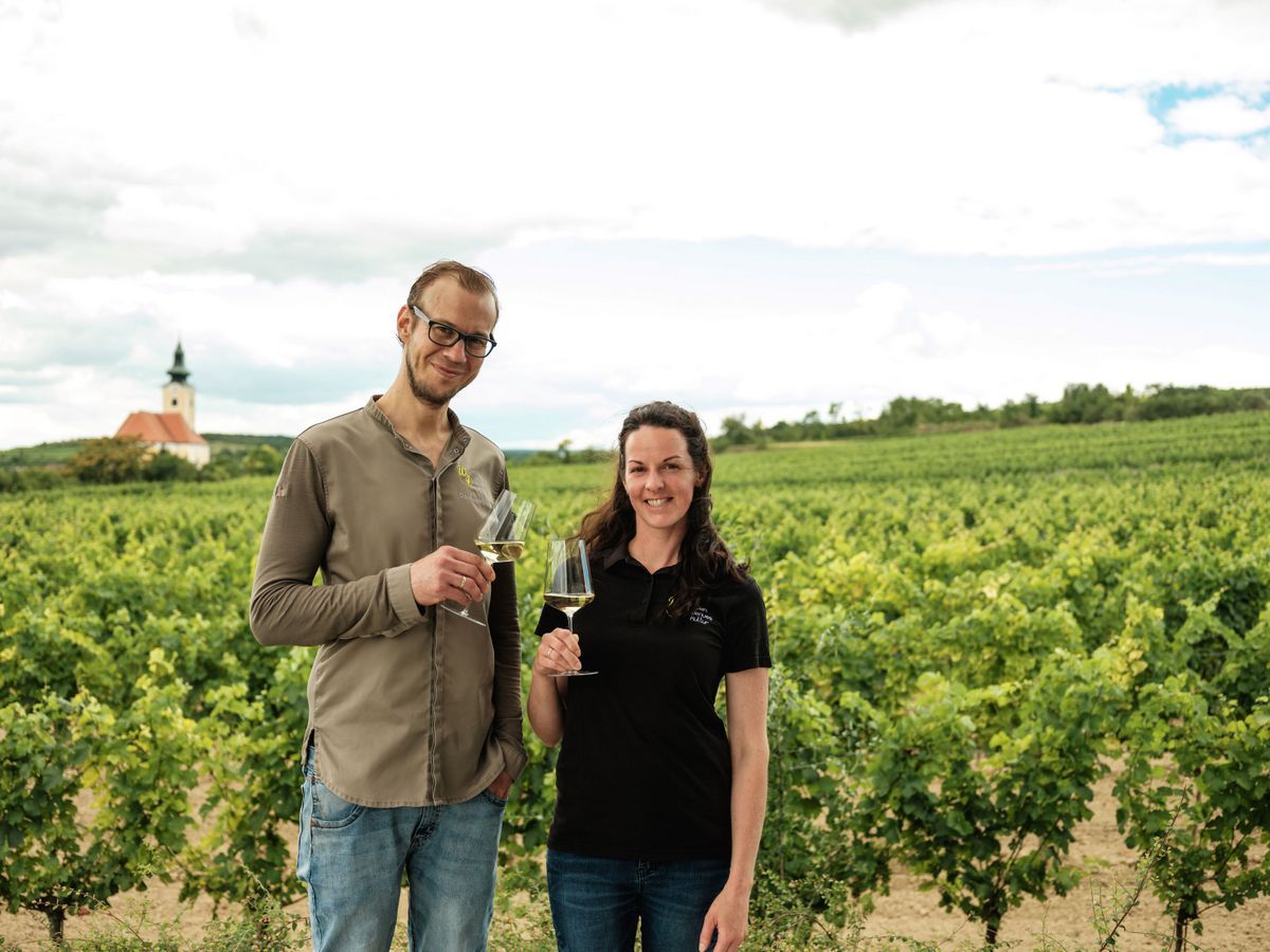 Two people stand with wine glasses in a vineyard, with a church in the background.