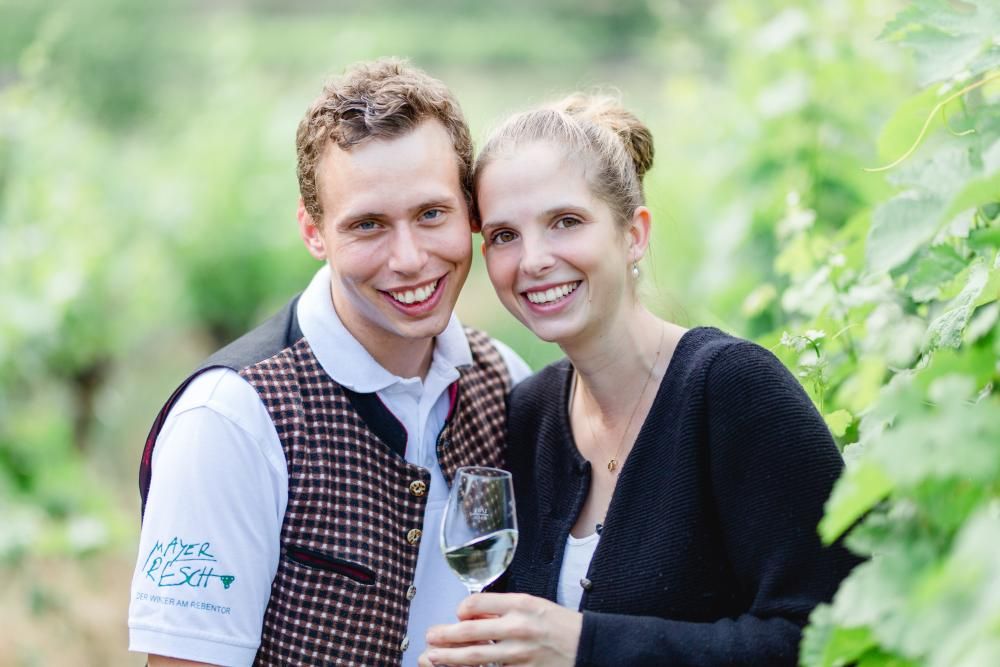 A smiling couple in a vineyard, the man wearing a vest, the woman holding a wine glass.