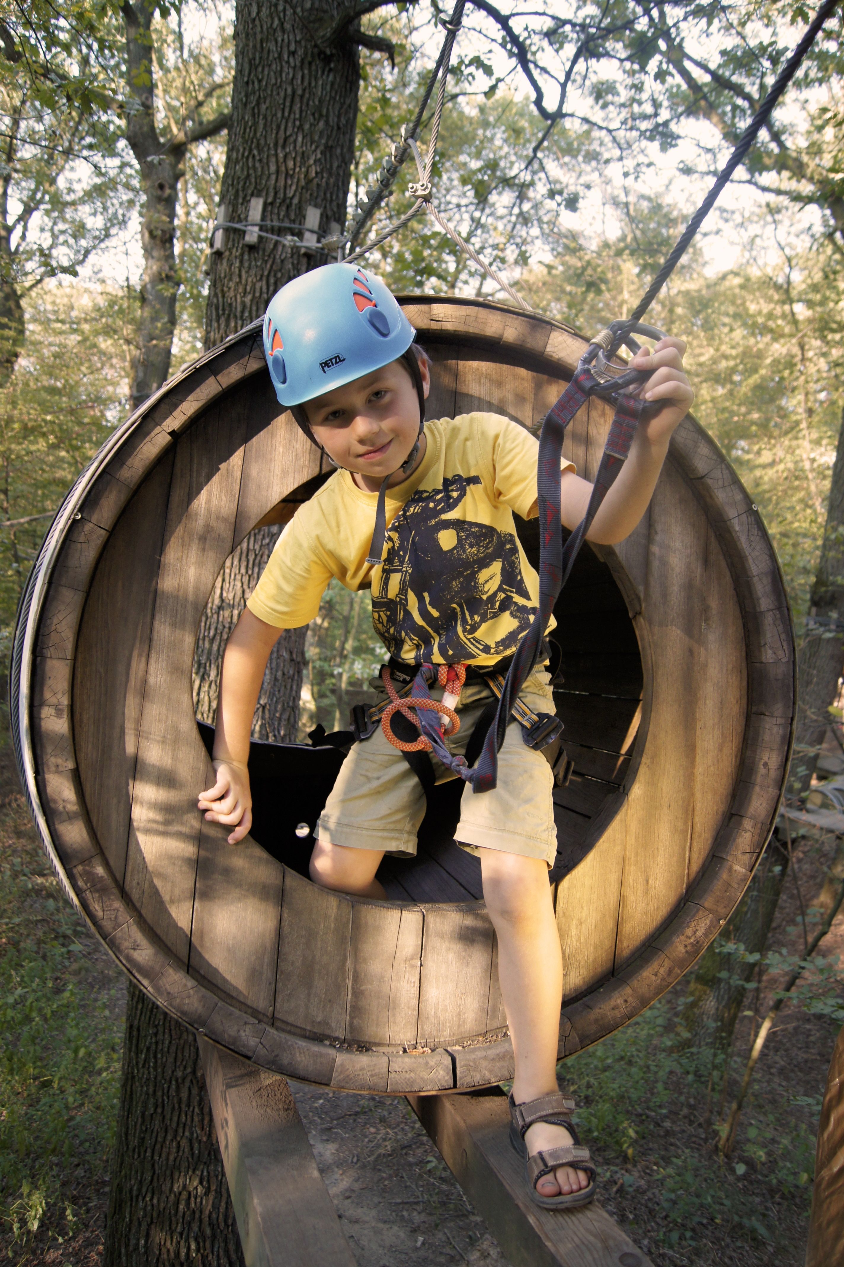 A child with helmet and climbing equipment in a wooden climbing park tunnel.