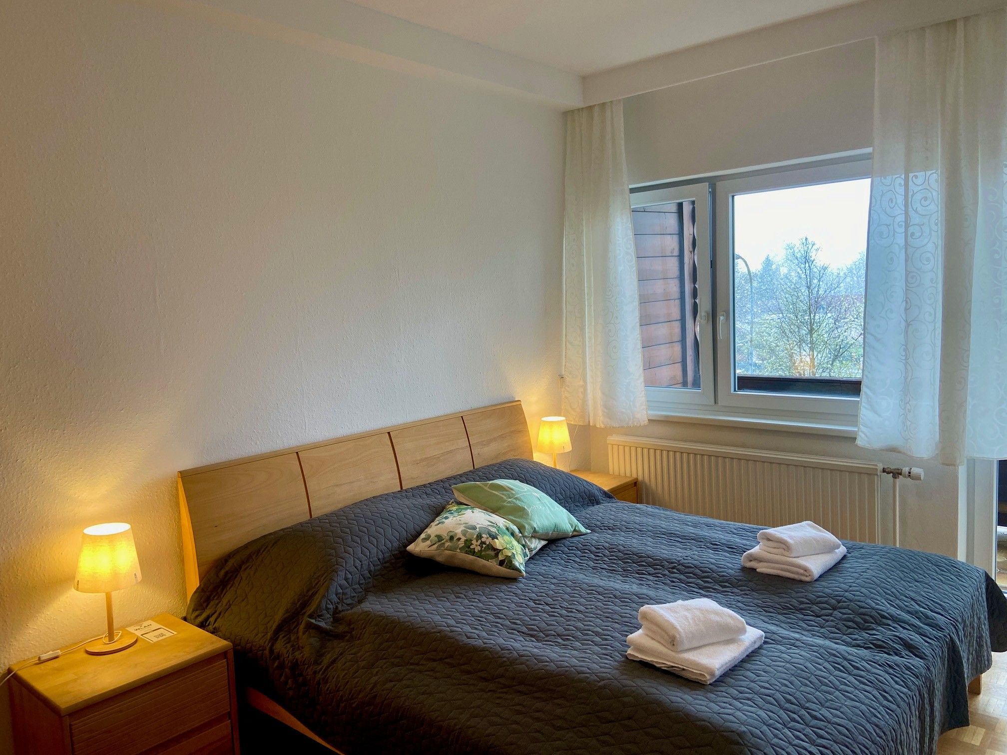 A cozy double room with a large bed, two bedside tables with lamps and a window with a view outside.