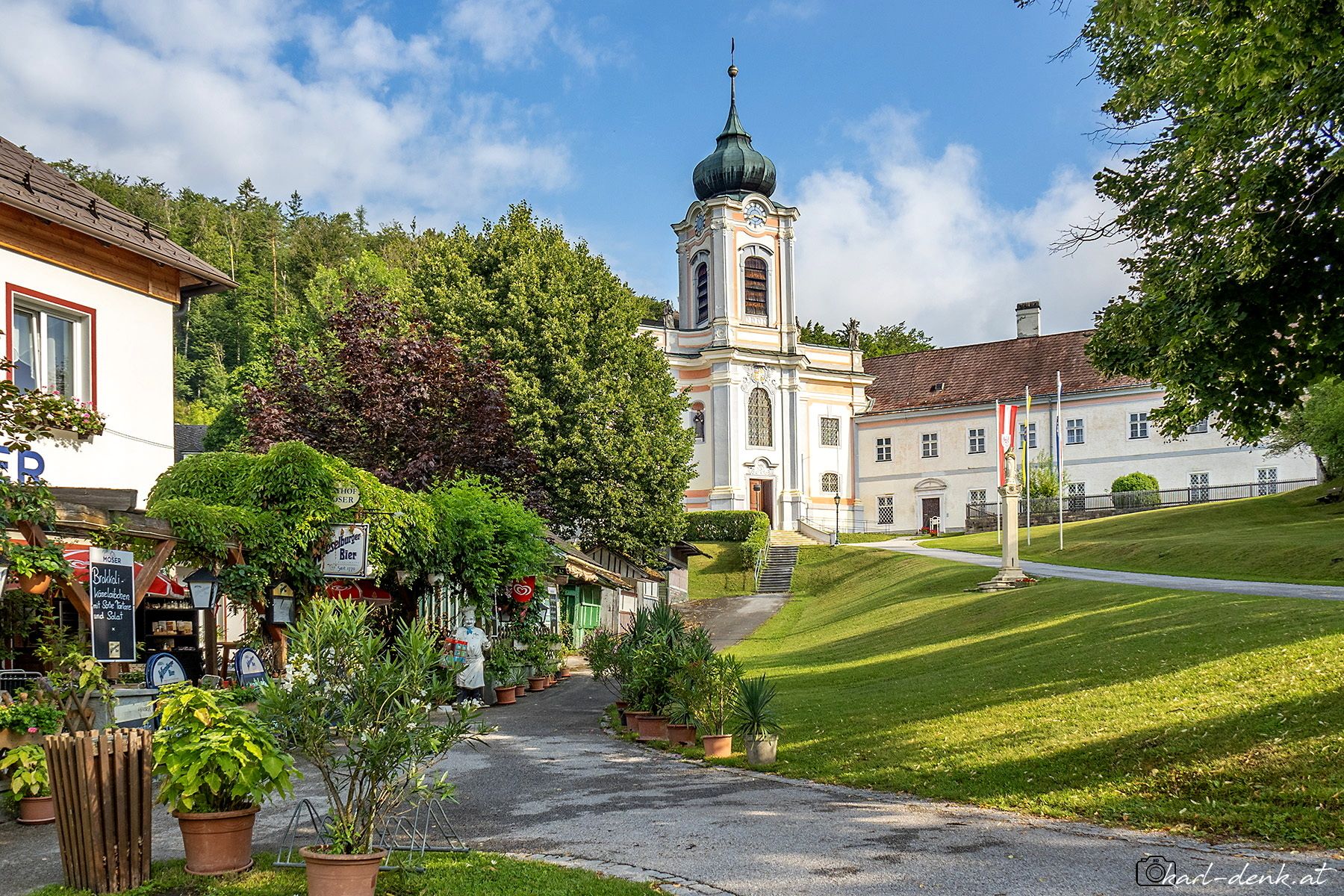A picturesque village with a Baroque-style church and an inn in the foreground, surrounded by green trees and a blue sky.