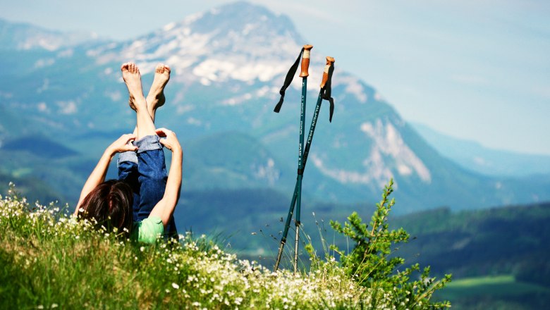 Person lying on a meadow with a view of the mountains, hiking poles next to them.