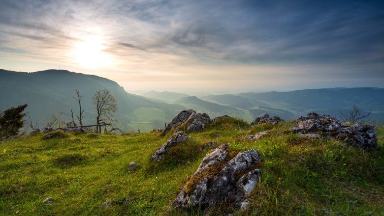 Panoramic view from a hill with rocks and grass in the foreground, mountains and a cloudy sky in the background.