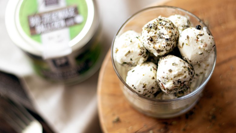 A glass filled with goat's cheese balls, sprinkled with herbs, on a round wooden board. In the background, another jar of goat's cheese balls with a green label.