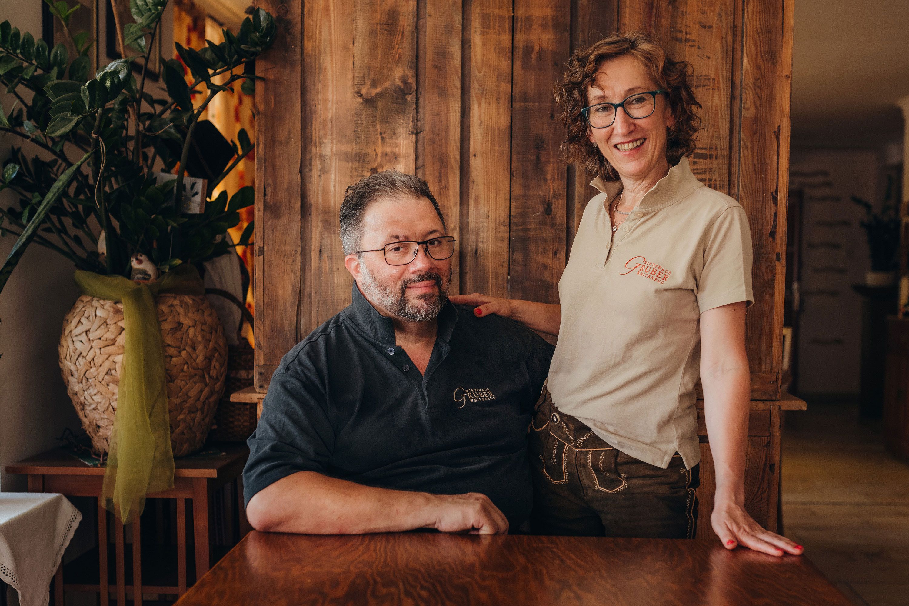 Two people in a rustic room, one sitting, the other standing with a smile.