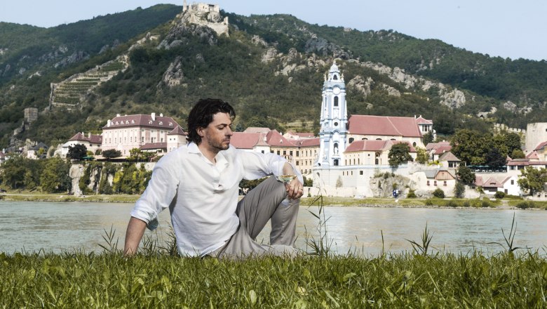 A man sits on a meadow against the backdrop of Dürnstein with the Danube in the background.