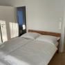 One bedroom with a double bed, white comforter covers and wooden floor. An open door leads to a bathroom.