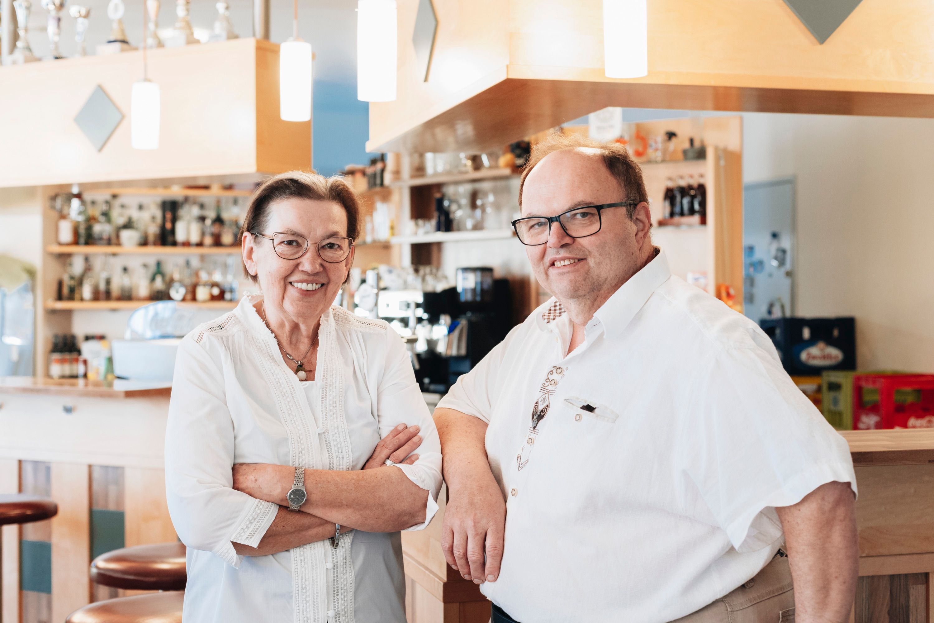 Two people in a café or bar, smiling, in front of a counter with bottles in the background.