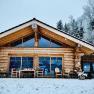 Log cabin in the snow with large windows and terrace, surrounded by snow-covered trees.