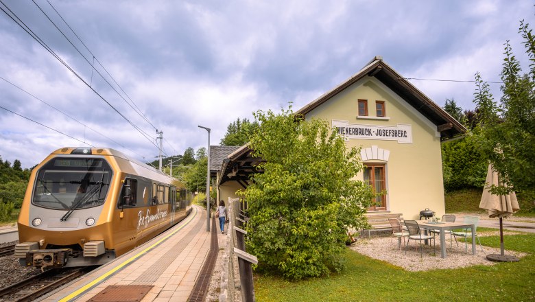 A golden train stops at Wienerbruck-Josefsberg station, surrounded by green countryside and a small outdoor seating area.