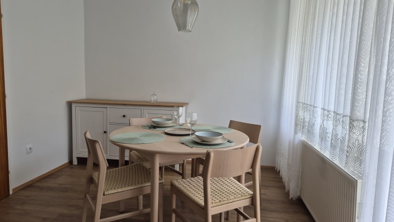 Dining area with round table, four chairs and crockery, next to a window with white curtains.