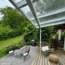 Terrace with glass roof, garden furniture and barbecue, surrounded by green vegetation.