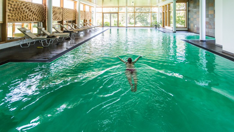 Person swimming in an indoor pool with green water and deckchairs at the edge.