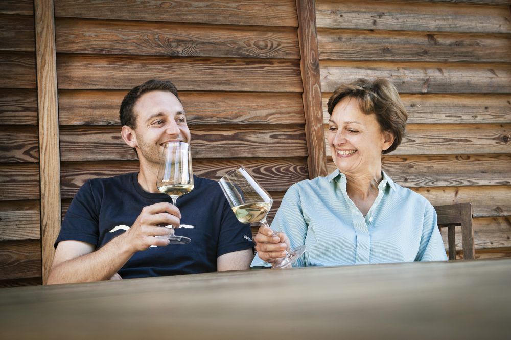 Two people clink glasses of wine, sitting in front of a wooden wall.