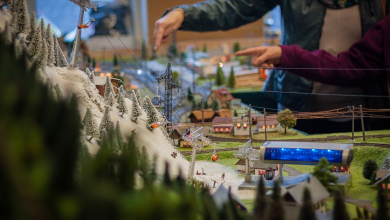 Detailed model landscape with cable car and snow-covered trees, two people pointing at the model in the background.