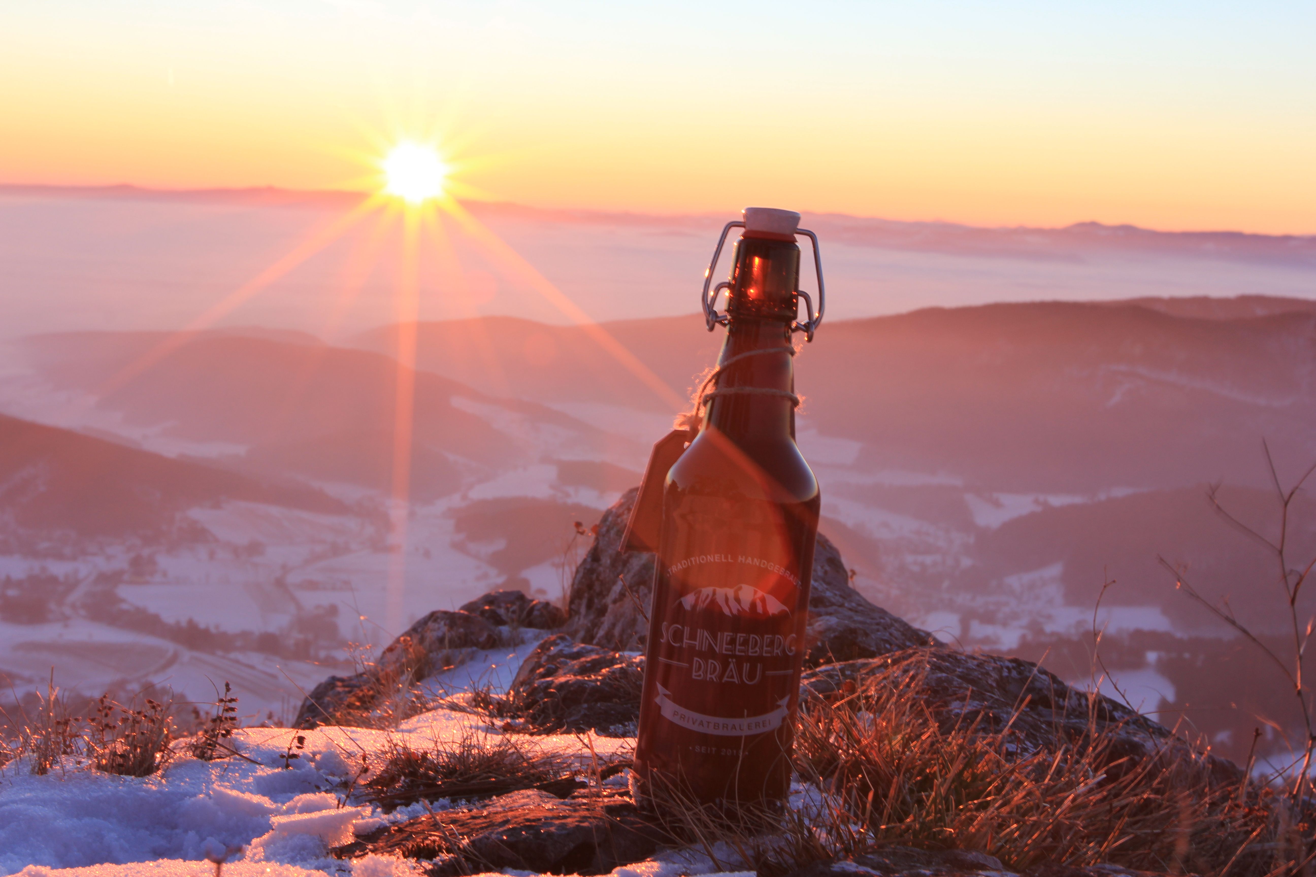 A bottle of Schneeberg Bräu beer stands on a snow-covered mountain at sunset.