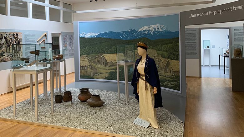 Exhibition in the Pitten Regional Museum with historical artifacts and a figure in traditional dress in front of a mural.