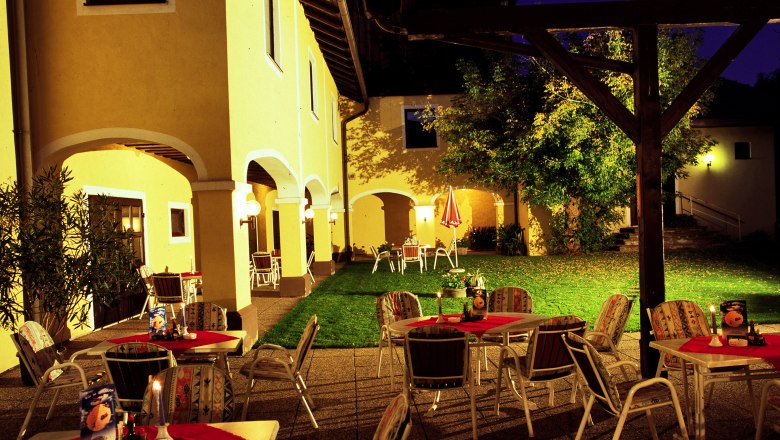 Gasthof Huber, © MG Rfd Illuminated outdoor area of an inn with yellow walls, tables and chairs on a terrace at night.