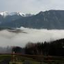 View of snow-covered mountains above a valley with fog and a wooden fence in the foreground.