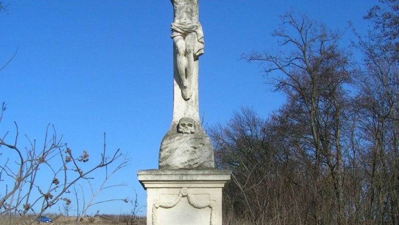 Stone cross with a figure of Jesus in an overgrown landscape under a blue sky.