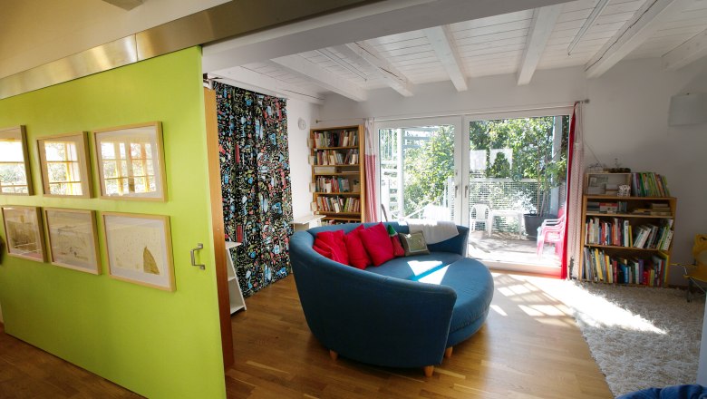 Interior view of a modern living room with a round blue couch, wooden floor, bookshelves and large windows overlooking a garden.