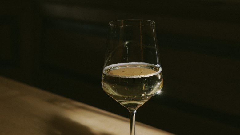 A wine glass with white wine stands on a wooden table in the shade.