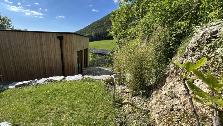 A modern tiny house with wooden cladding stands in a green, hilly landscape. Rocks and plants can be seen in the foreground.
