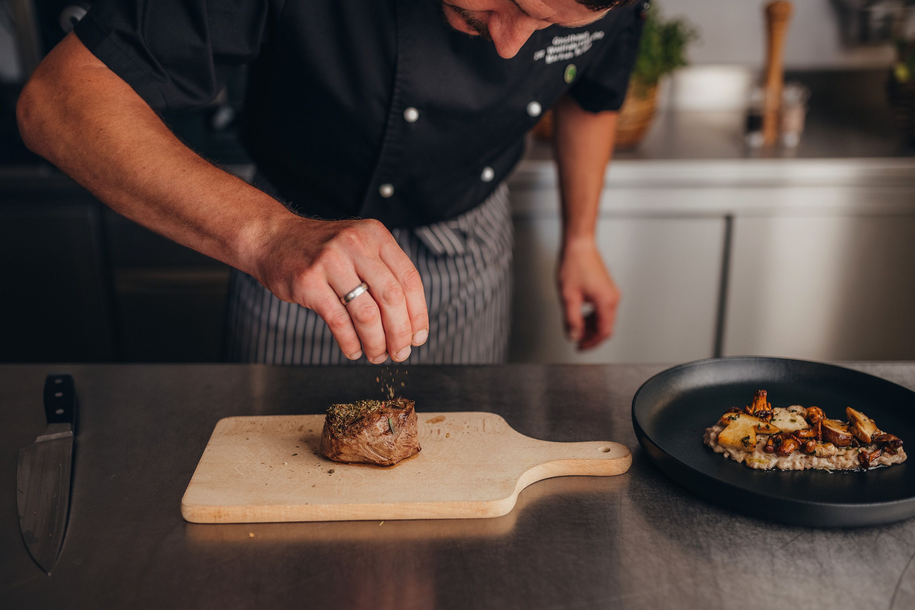 A cook seasons a piece of meat on a wooden board in a kitchen.
