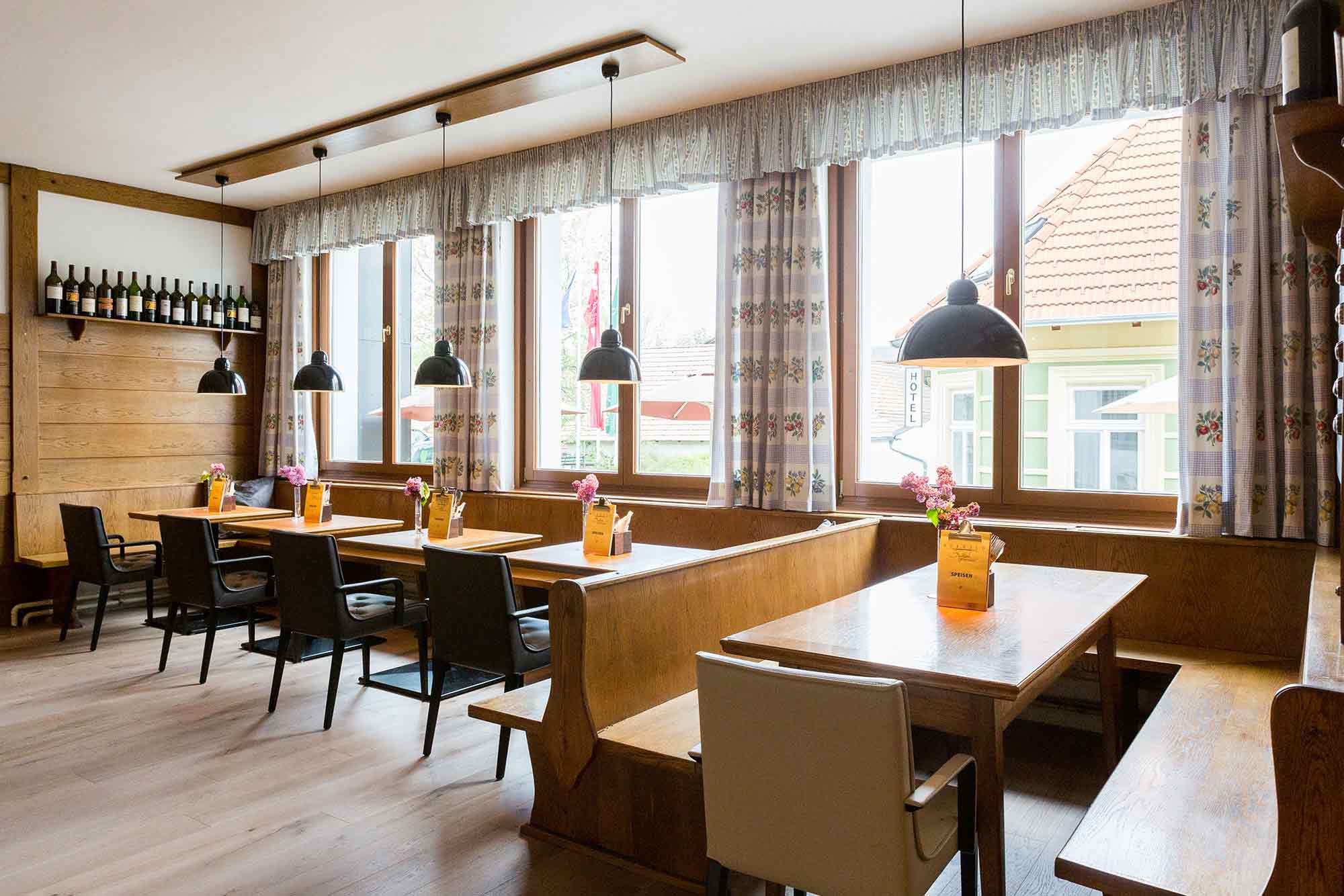 Cozy guest room with wooden tables, chairs and benches, decorated with flowers and menus. Large windows with curtains and hanging lamps.