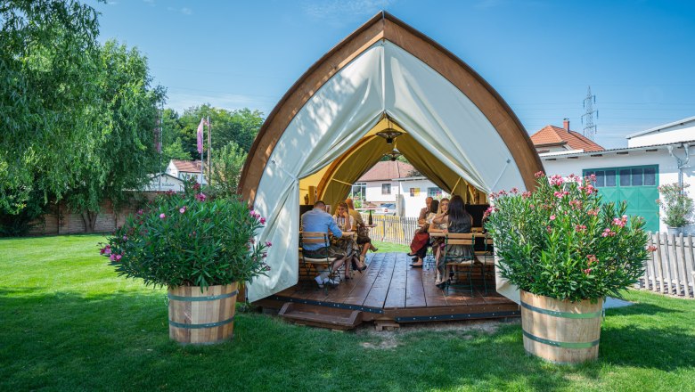 A tent with a wooden structure on a meadow where people are sitting at tables. In front of the tent are large planters with flowers.