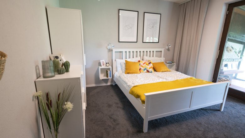 Bedroom with white bed, yellow pillows and blanket, two murals and a window with curtain.
