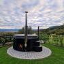 A round hot tub with an outdoor fireplace, surrounded by green countryside and cloudy skies.
