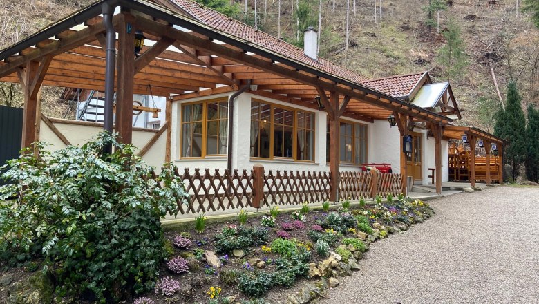 An inn with a wooden roof and flower garden in front of a wooded slope.