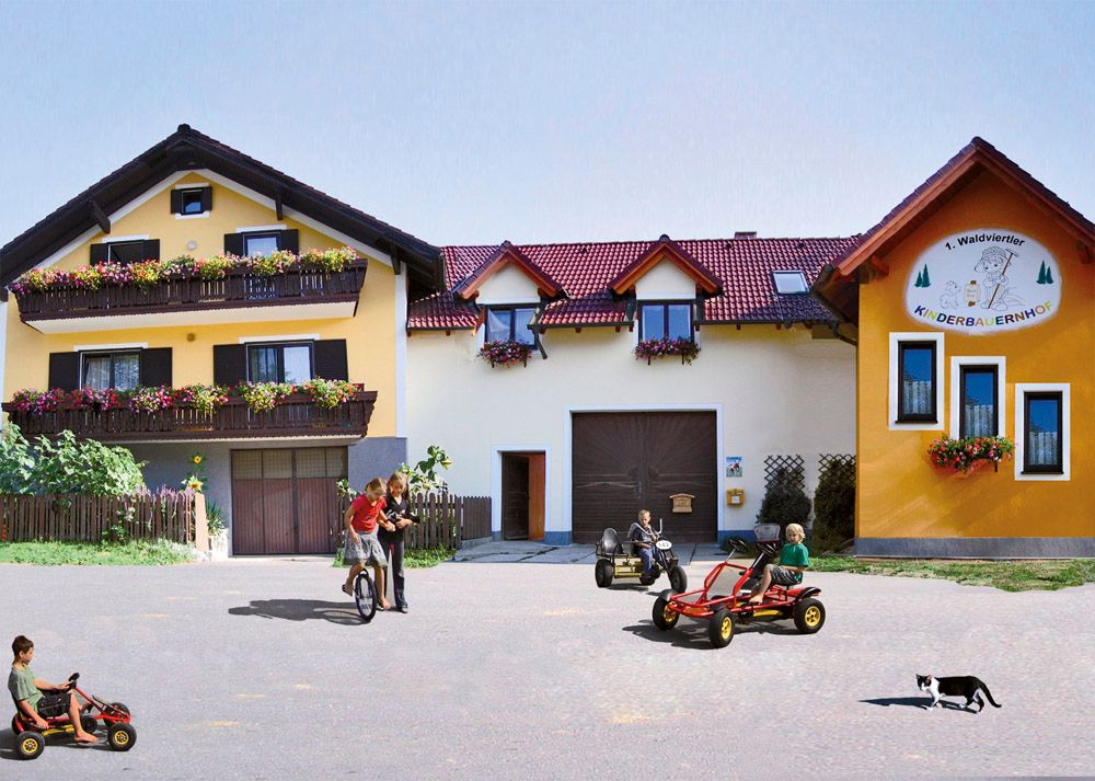 Children play on Kettcars in front of a yellow farm with flower balconies.
