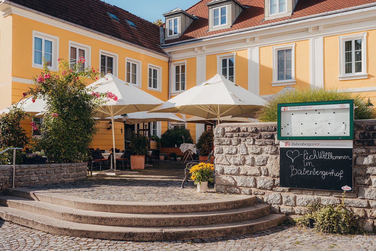 Yellow historic building with terrace and parasols, surrounded by plants.