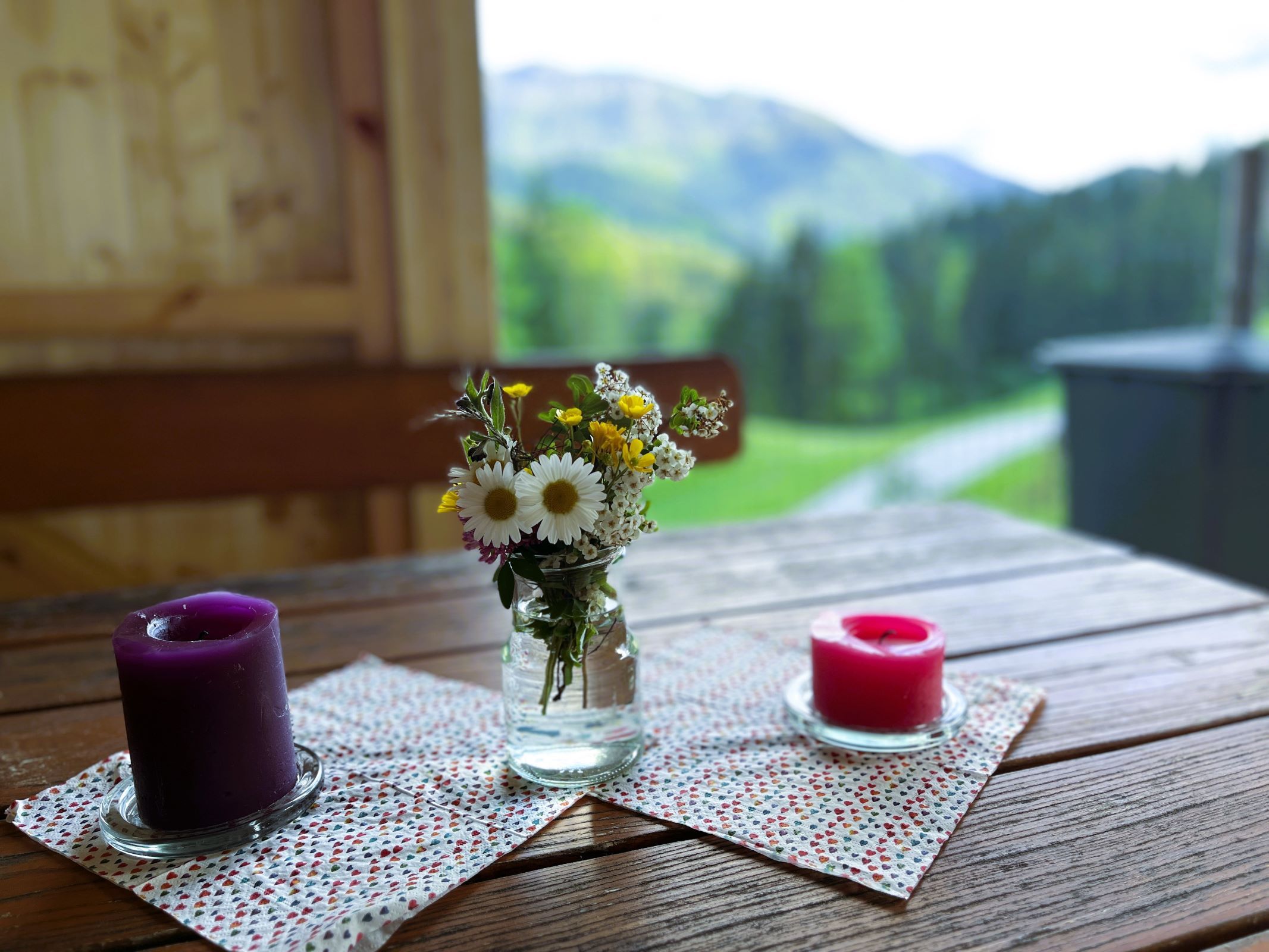 A wooden table with two candles and a small bouquet of flowers in a glass jar, a blurred mountain landscape in the background.