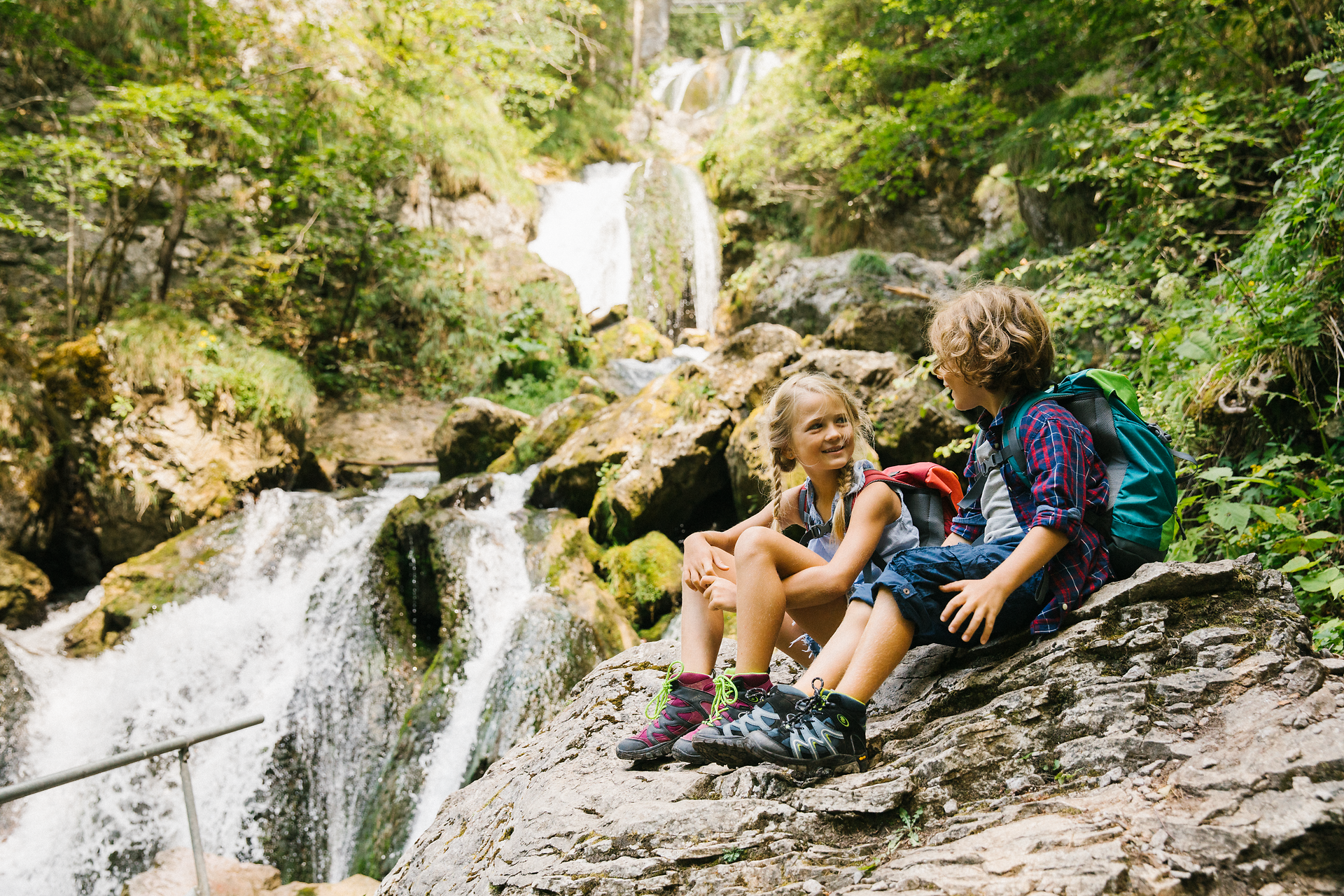 Two children sit relaxed on a large stone and enjoy the refreshing atmosphere of the nature park. Surrounded by lush greenery and the gentle sound of the water, the scene radiates pure joie de vivre and a sense of adventure.