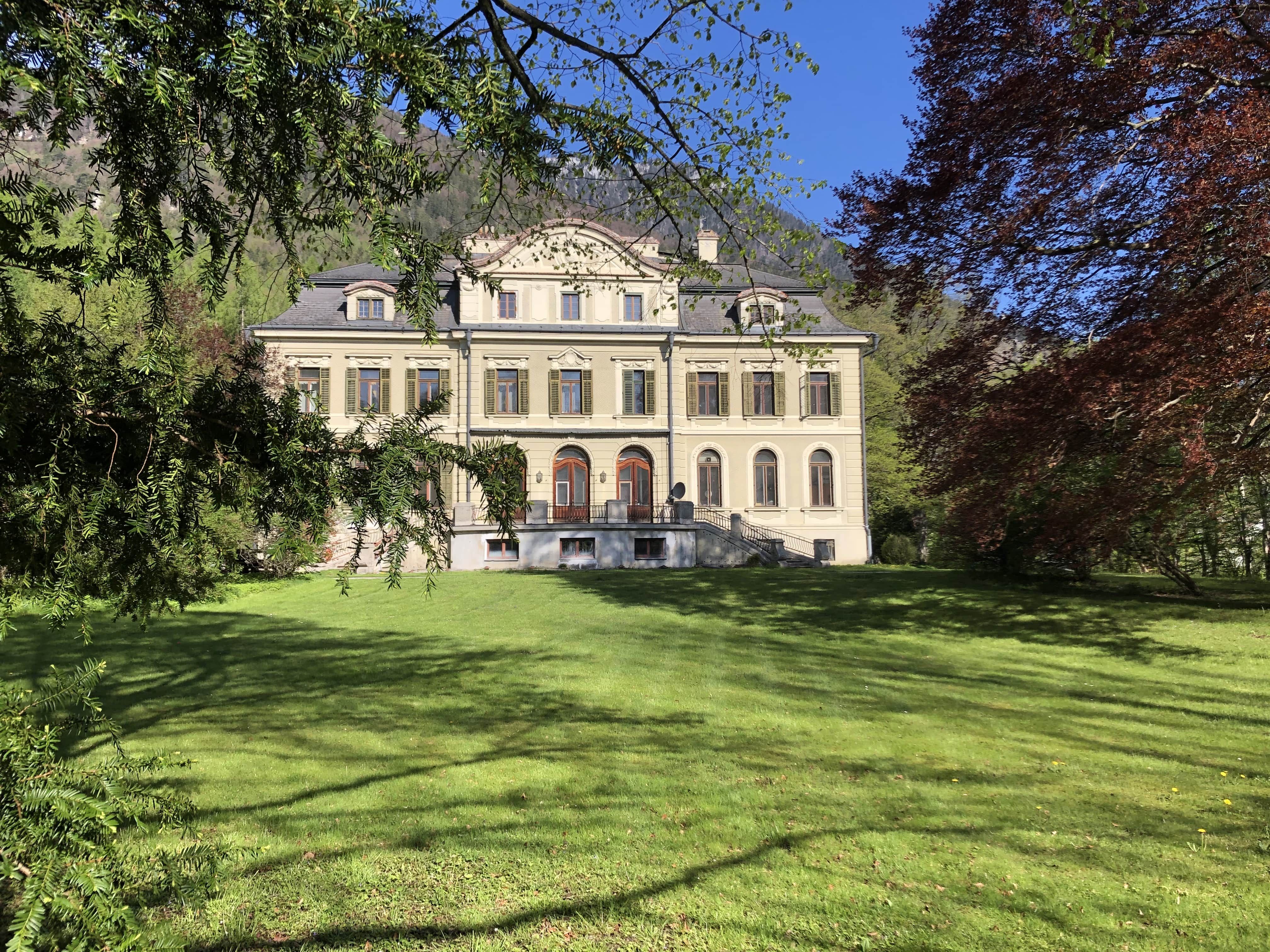 A large, elegant manor house with a symmetrical façade and several windows, surrounded by green lawns and trees.