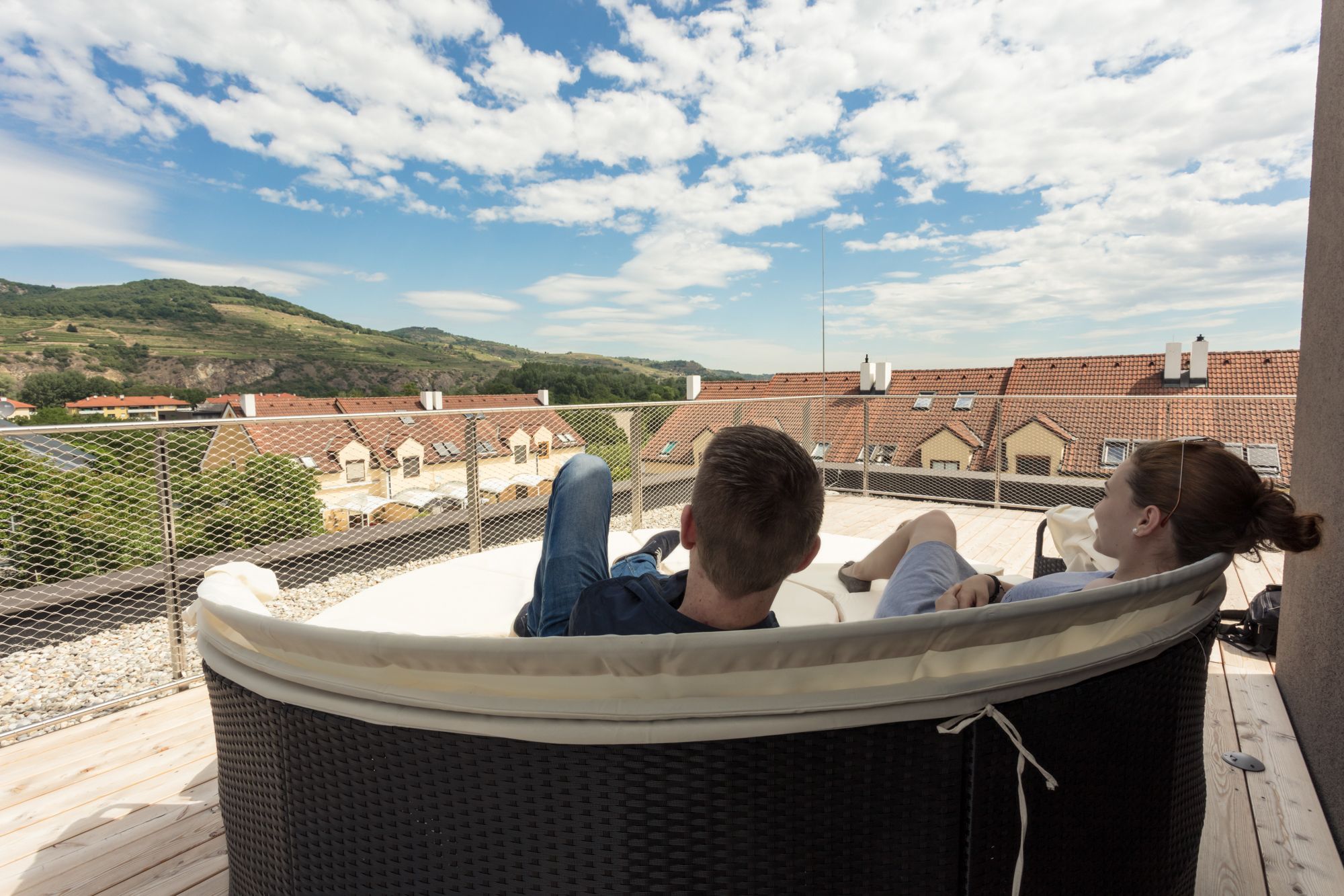 Two people relax on a terrace with a view of rooftops and hills.