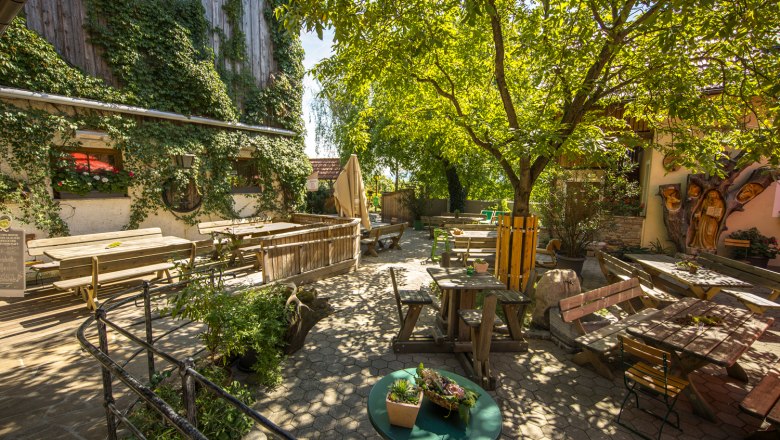 A cozy guest garden with wooden tables and benches under a large tree, surrounded by ivy-covered walls.