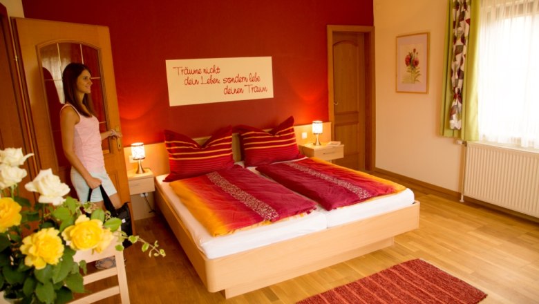 A cozy bedroom with a red wall, double bed and flowers. A woman enters the room.