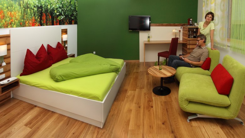 A modern guest room with a green bed, sofa and wooden floor. There are two people in the room, one sitting on the sofa, the other standing next to it.