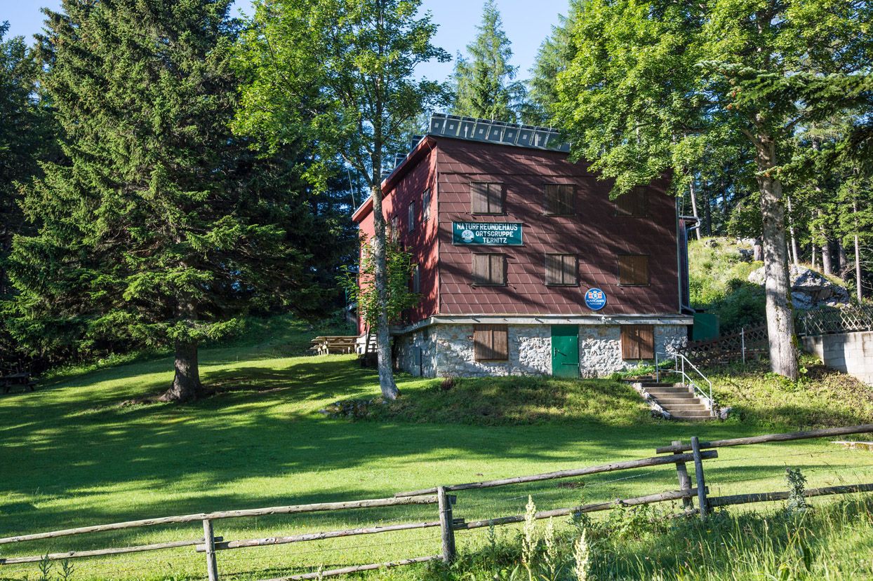 A mountain hut surrounded by trees and meadows, with a wooden fence in the foreground.