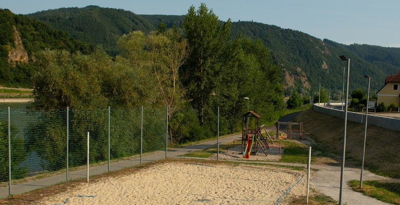A beach volleyball court next to the Danube with surrounding trees and a playground in the background.