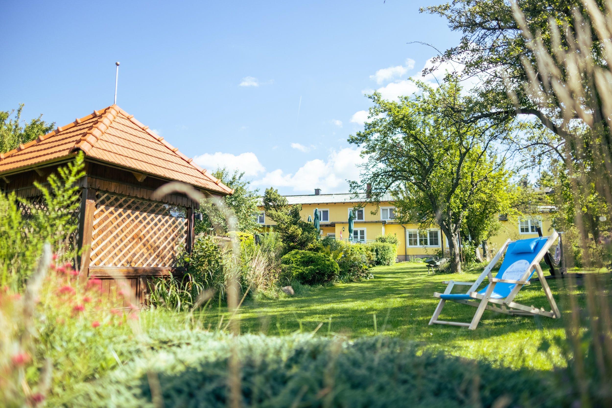 A green garden with a pavilion, deckchairs and a yellow building in the background.