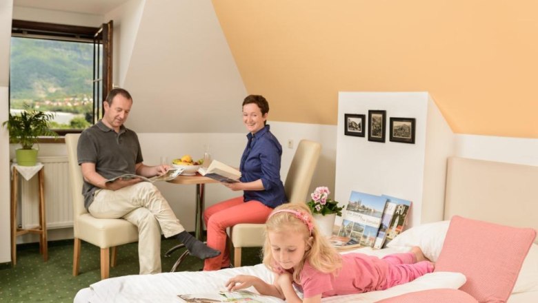 Family in a cozy room, parents reading at the table, child lying on the bed with a book.