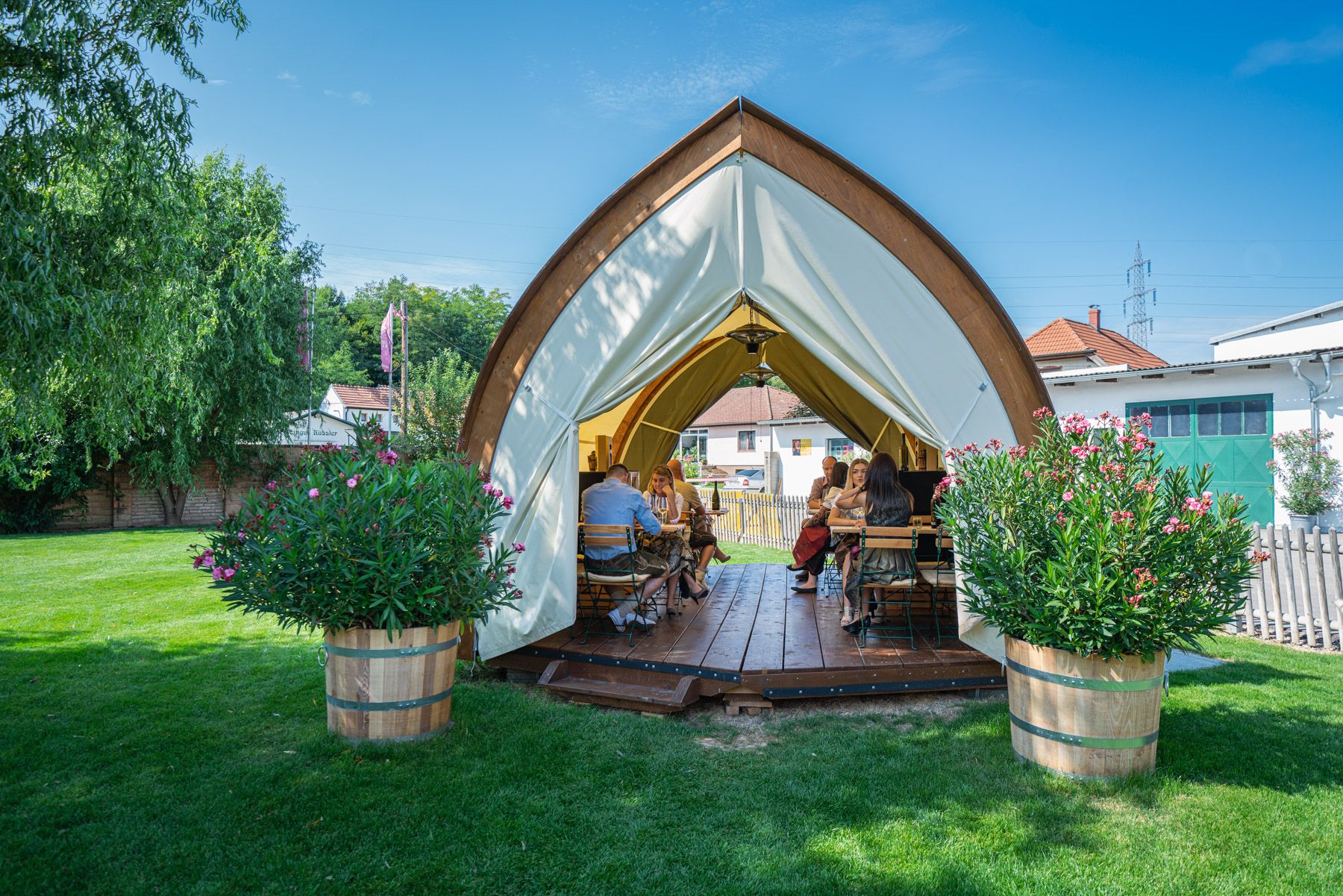 A tent with a wooden structure on a meadow where people are sitting at tables. In front of the tent are large planters with flowers.